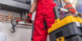 Technician in red overalls holding a toolbox in a plumbing setting, emphasizing HVAC repair and installation services.