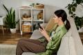 Woman sitting on a sofa in a cozy living room, using a smartphone, surrounded by indoor plants and home decor, emphasizing comfort and relaxation in a well-maintained home environment.