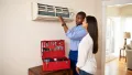 Technician explaining air conditioning unit repair to a homeowner, with a toolbox of tools in the foreground, emphasizing emergency HVAC services in a residential setting.