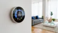 Smart thermostat mounted on a wall, displaying temperature settings, in a modern living room with a gray sofa and decorative cushions.
