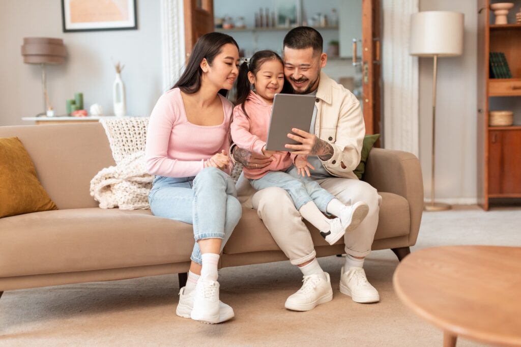 Korean family enjoying time together on a couch, watching cartoons on a tablet in a cozy living room, highlighting family comfort and quality time.