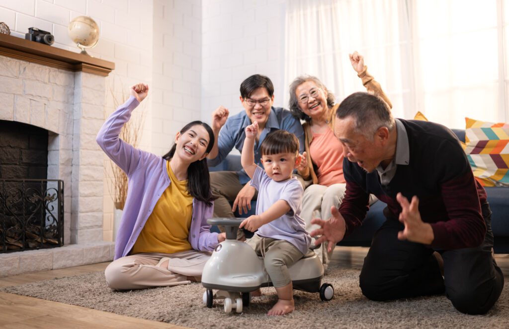 Happy multi-generational Asian family celebrating together in a cozy living room, with a child playing on a toy vehicle, emphasizing family warmth and joy.