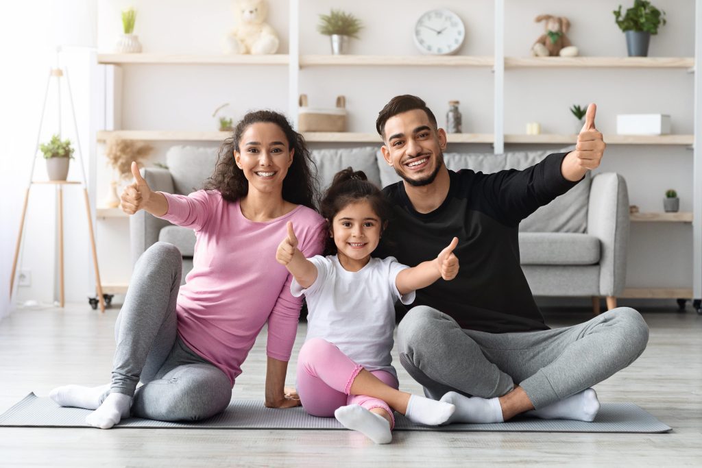 Family exercising together at home, smiling and giving thumbs up, promoting wellness and fitness.