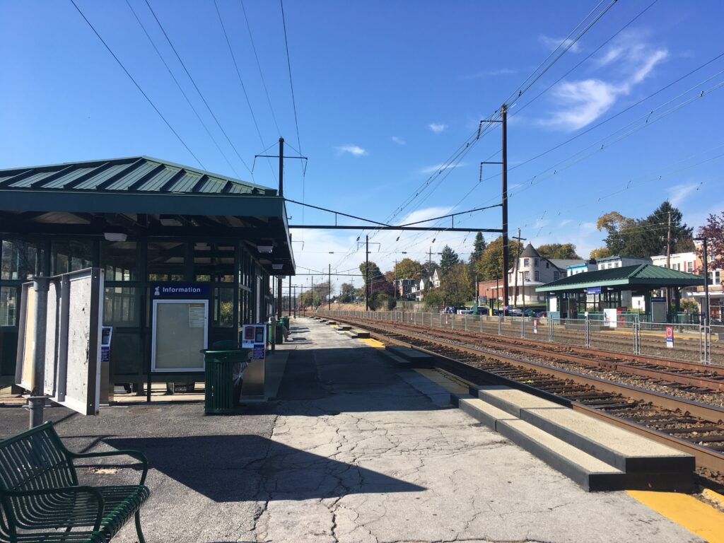 Downingtown: Train station platform view with tracks, shelters, and overhead lines under a clear blue sky. Public transportation scene.
